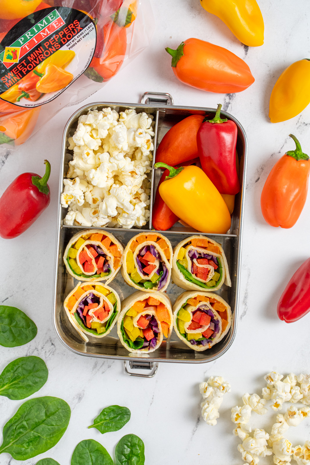 A bento box with veggie pinwheels, mini peppers, and popcorn.